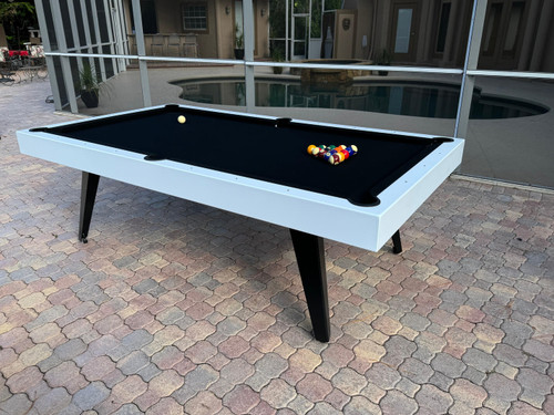A styled ¾ view of the Manhattan Outdoor Slate Pool Table, featuring a white aluminum frame and black pedestal legs, set in a modern outdoor lounge with mid-century accents.