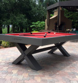 The Tasmania Outdoor Pool Table in a white finish, shown in a modern courtyard with clean stonework and lounge furniture. Features a one-piece slate playfield, Sunbrella felt, and cross-leg base designed for outdoor use.
