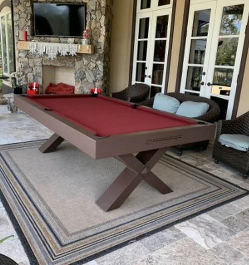 The Tasmania Outdoor Pool Table in a rich brown finish, placed beside a luxury pool with wood decking and palm trees. Built with a single-piece slate bed and finished with moisture-resistant Sunbrella felt for all-season play.